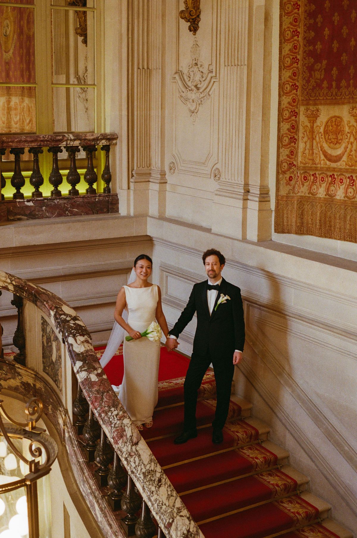 The bride and groom stand holding hands on a staircase with a red carpet and gilded balustrade, seen from above.