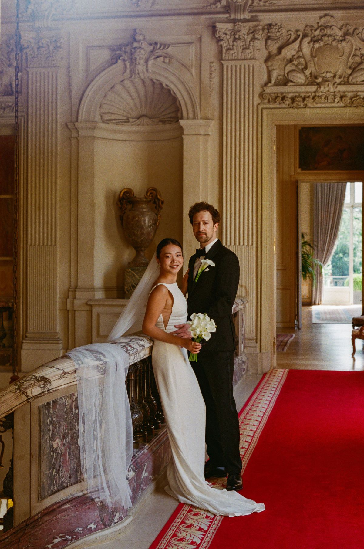 The couple stand close together beside a stone balustrade and red carpet in a warmly lit, ornate hallway, the bride holding flowers.