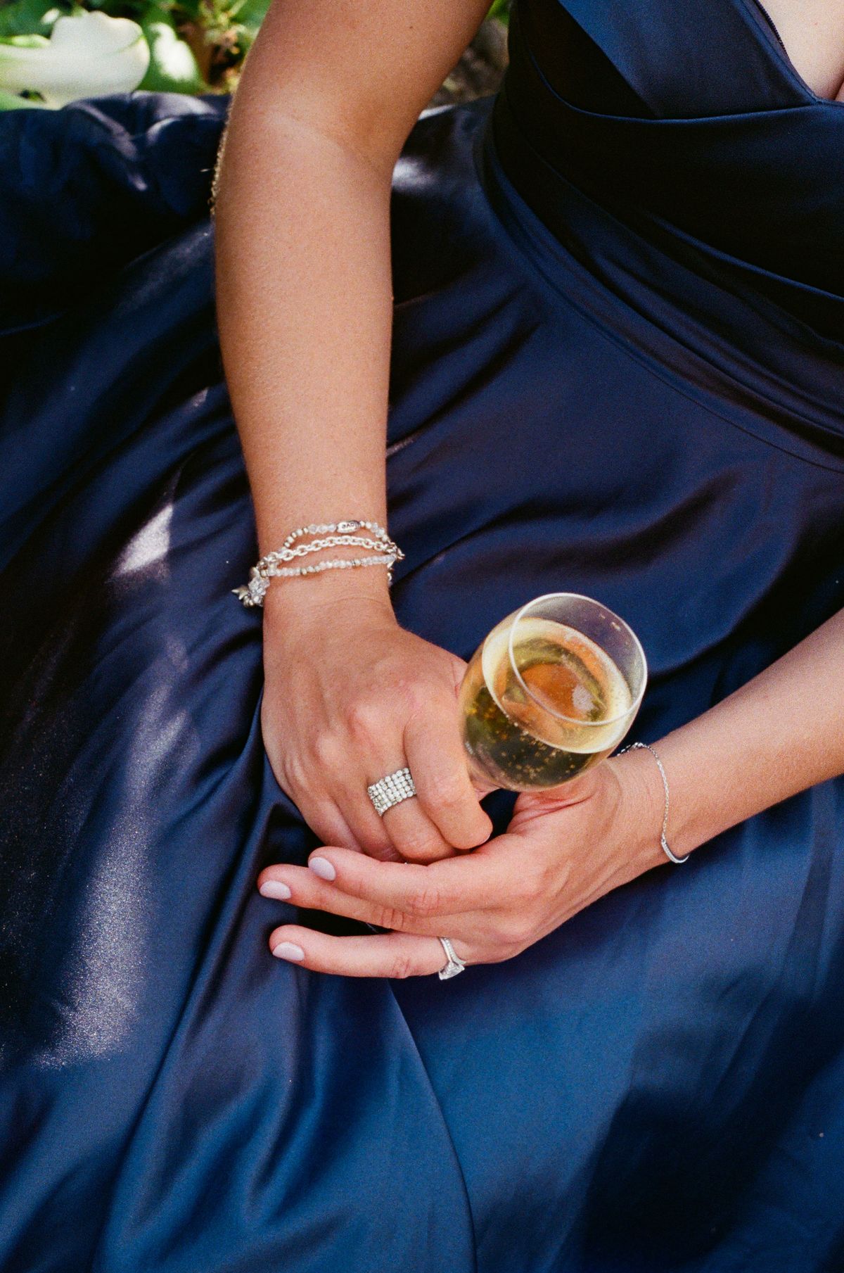 A woman in a navy satin dress, wearing a bracelet and rings, holds a glass of champagne in her lap.