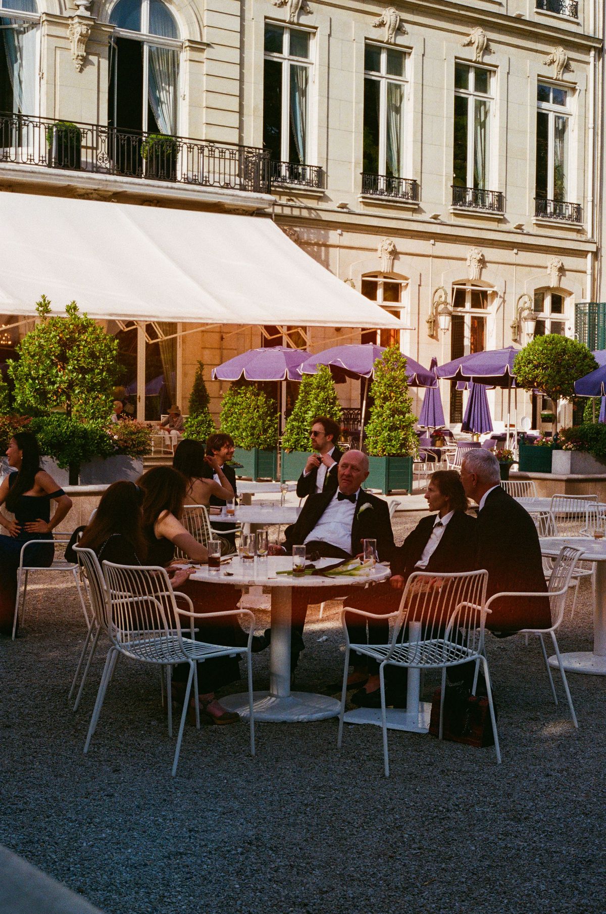 Guests in formal clothes sit around round tables on a gravel terrace beneath a white awning, with purple parasols and a stone building behind.