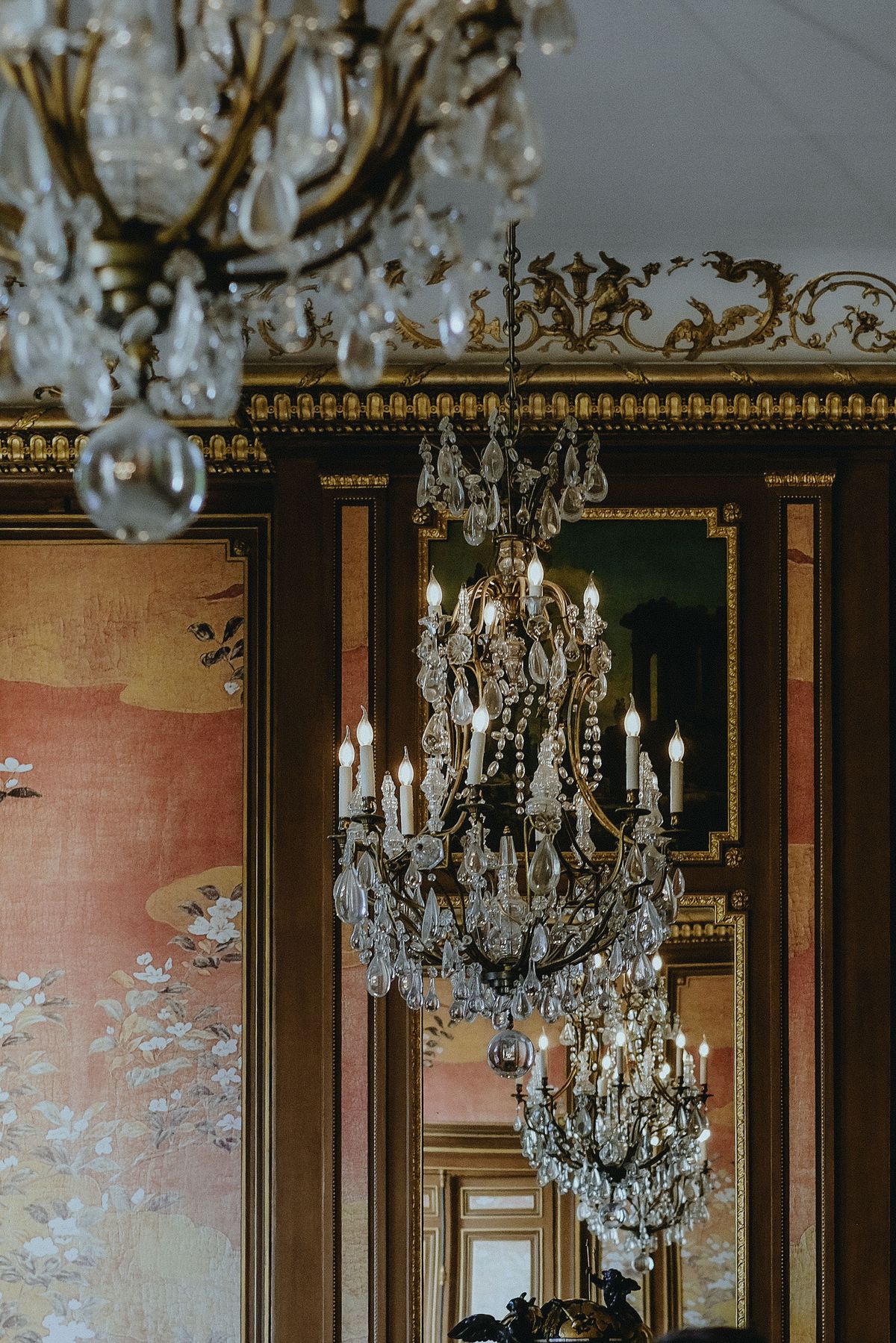 Crystal chandeliers hang in a salon with gilded mouldings and walls painted with soft pink and green motifs.