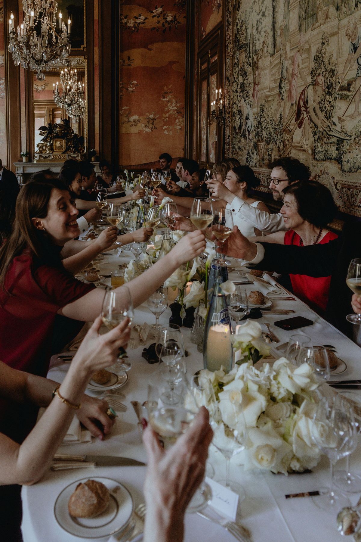 Guests seated along a long table raise glasses and applaud, with white flowers, candles and plates set between them.