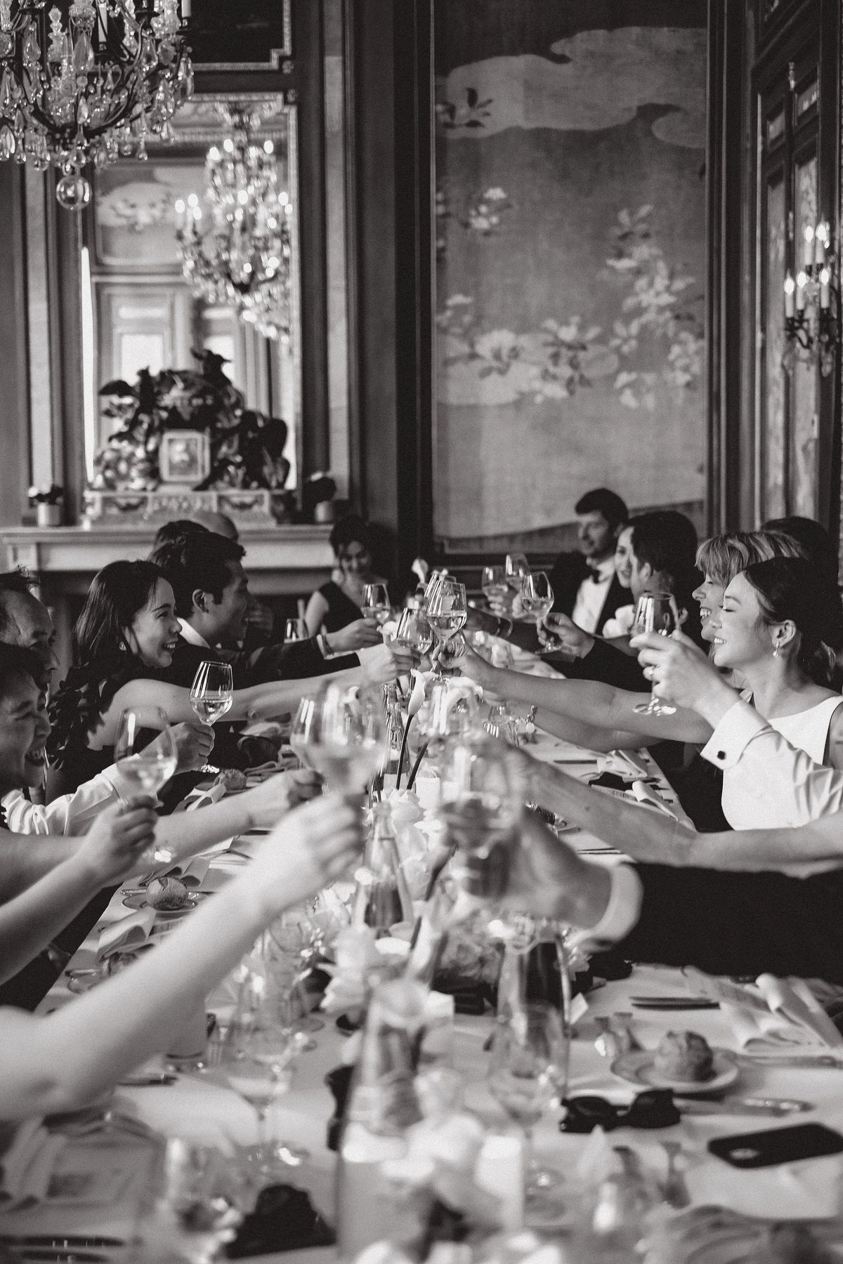Guests seated along a long table reach across with raised glasses to toast, among flowers and tableware, in black and white.