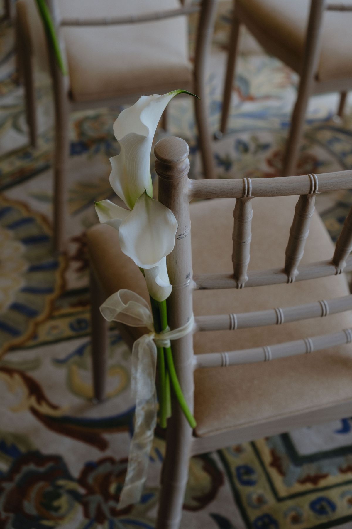 A white calla lily tied with ribbon to the back of a light wooden chair on a patterned carpet.