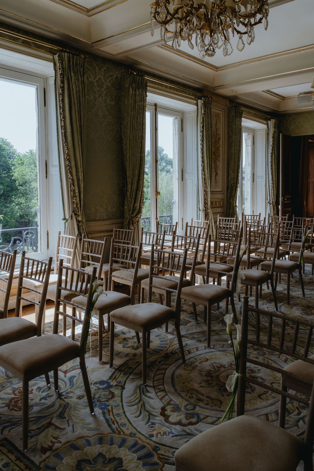 Rows of light wooden chairs face tall windows with green curtains in an elegant room with a gold chandelier.