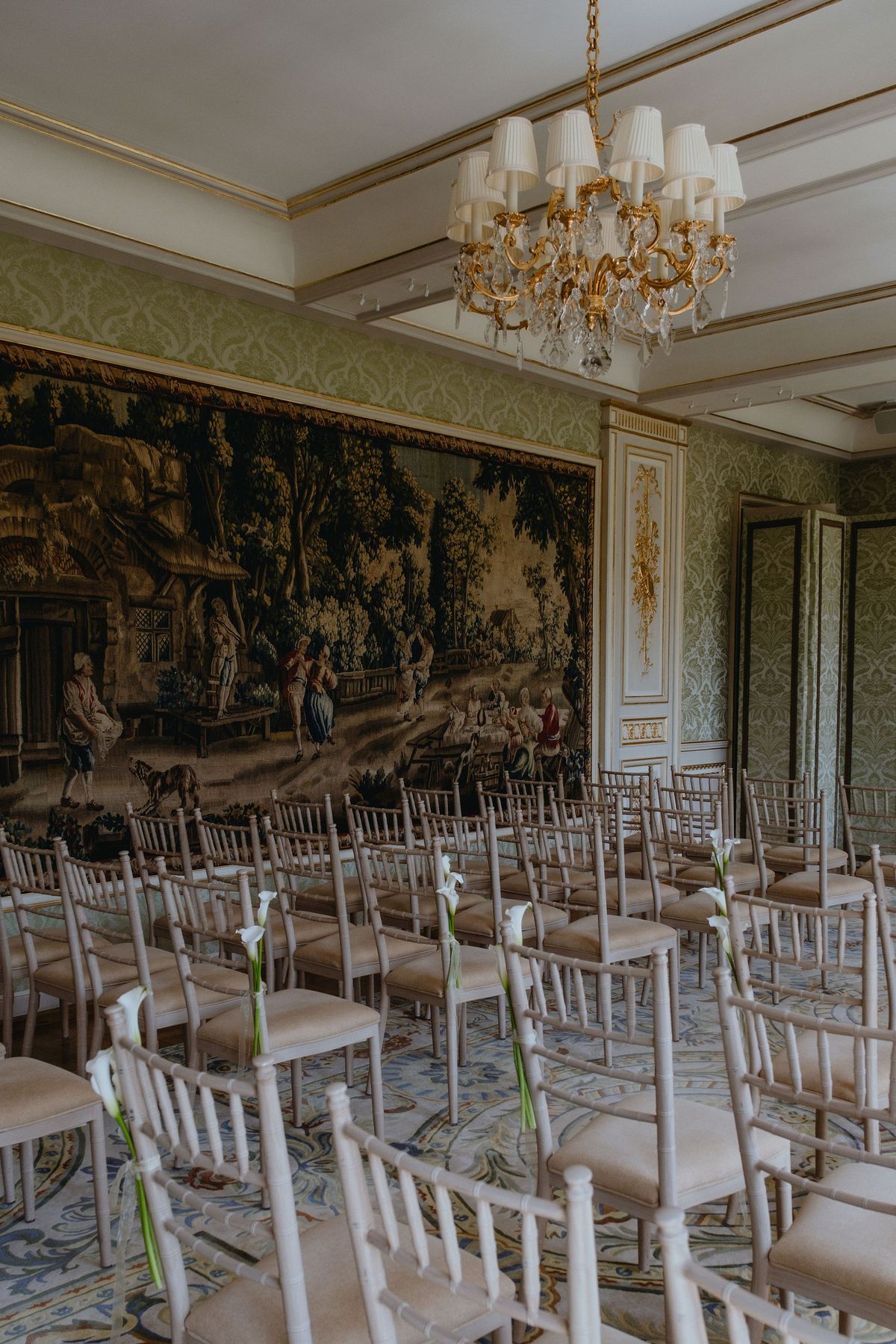 Rows of white chairs face a large tapestry in a room with a gold chandelier and pale green walls.