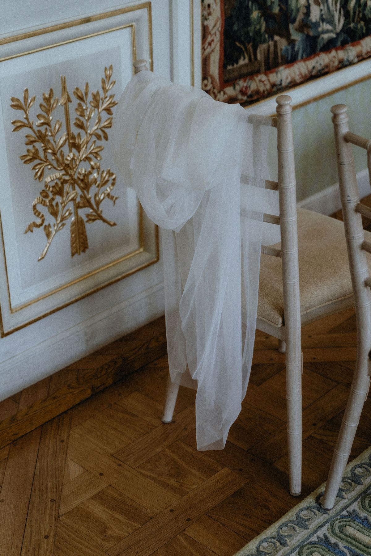 A white wooden chair is tied with soft white tulle, standing on a parquet floor beside a gilded panelled wall.