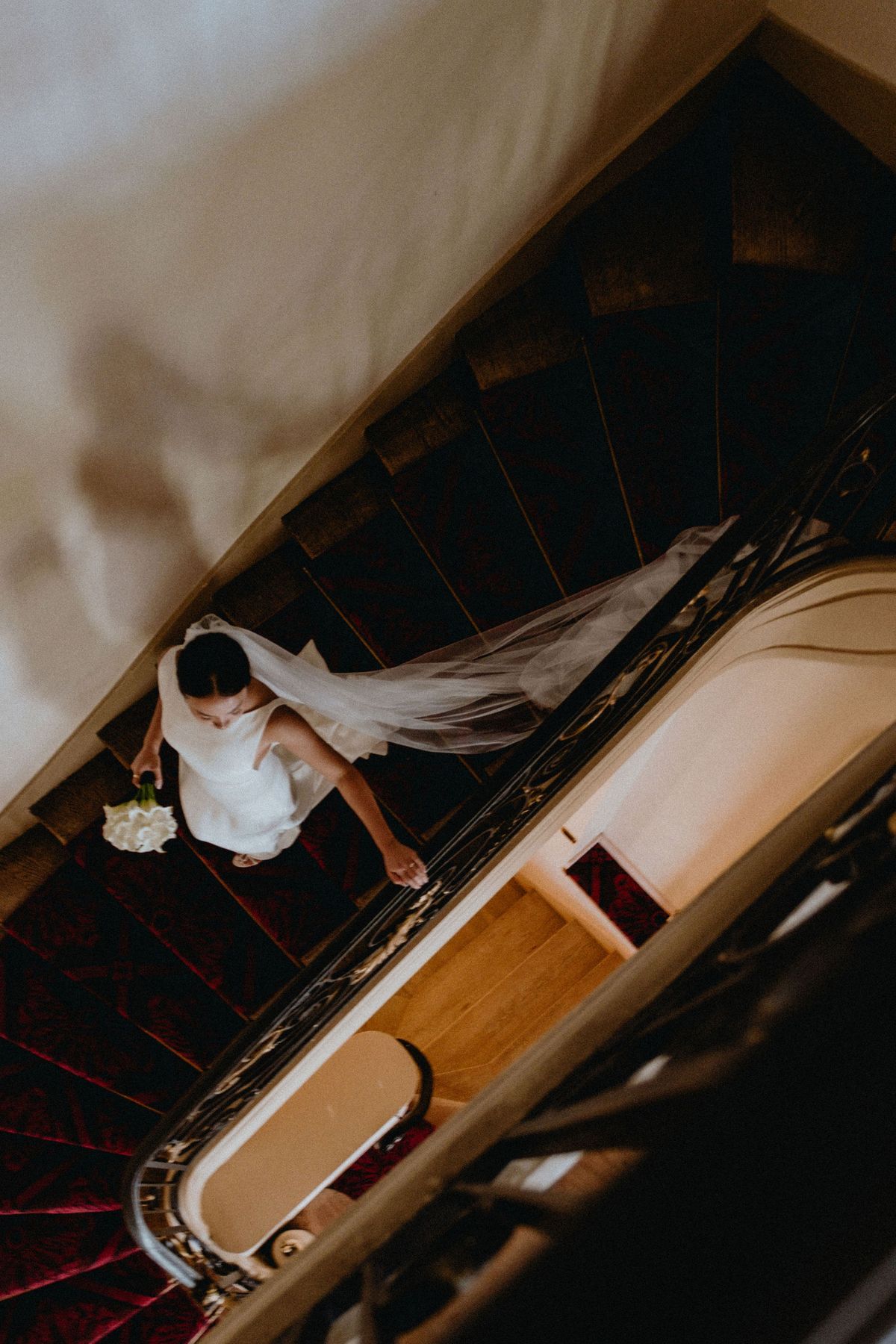 Seen from directly above, the bride in a white dress and veil descends a dark staircase with a curved wooden handrail.