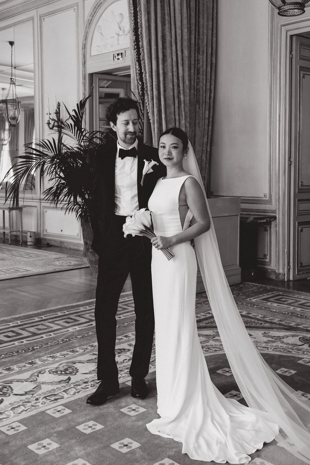 In black and white, the groom in a tuxedo and the bride in a long veil stand together in a grand room.