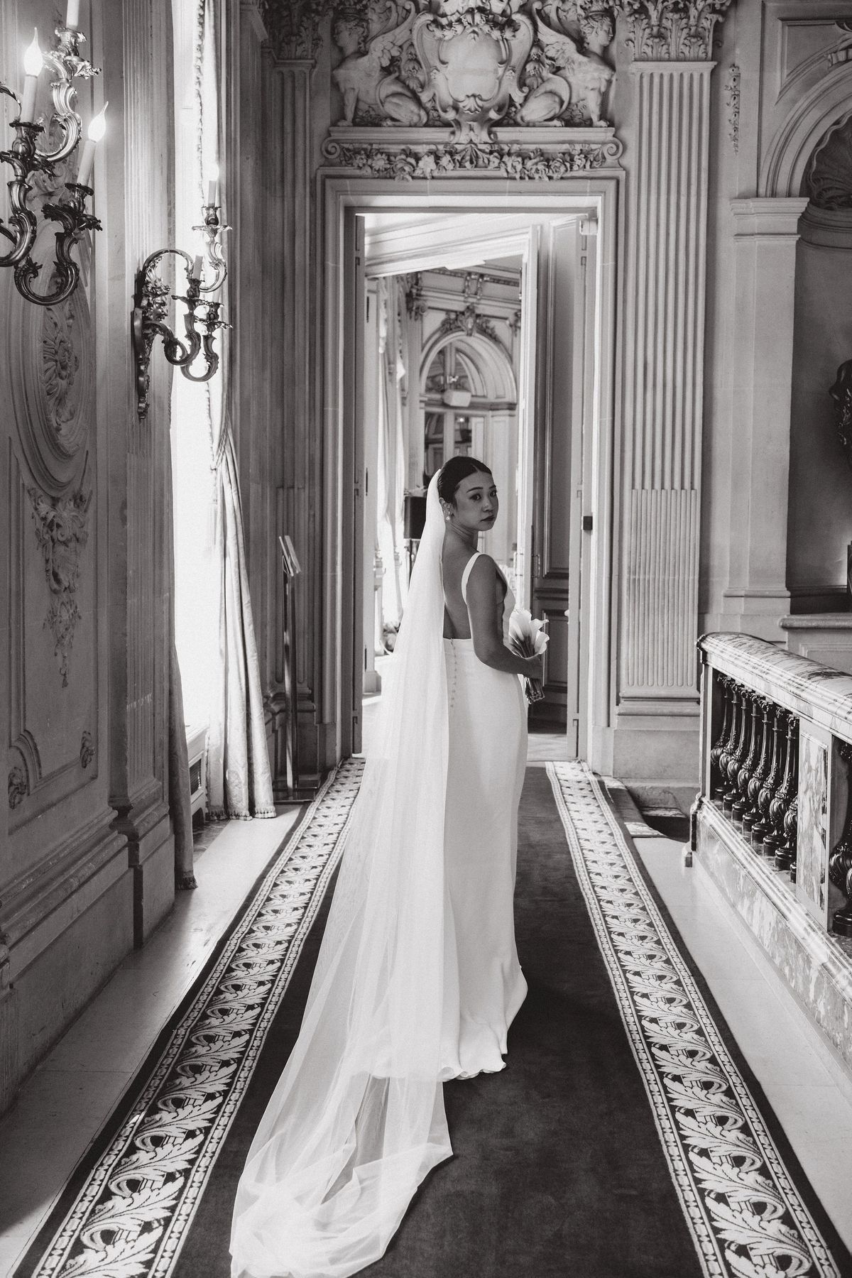 In black and white, the bride stands in a long ornate corridor with her veil trailing along a patterned runner.