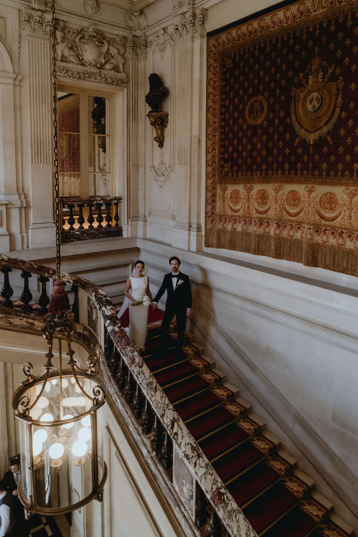 Seen from above, the couple stand on a red-carpeted marble staircase beneath ornate walls and a hanging lantern.