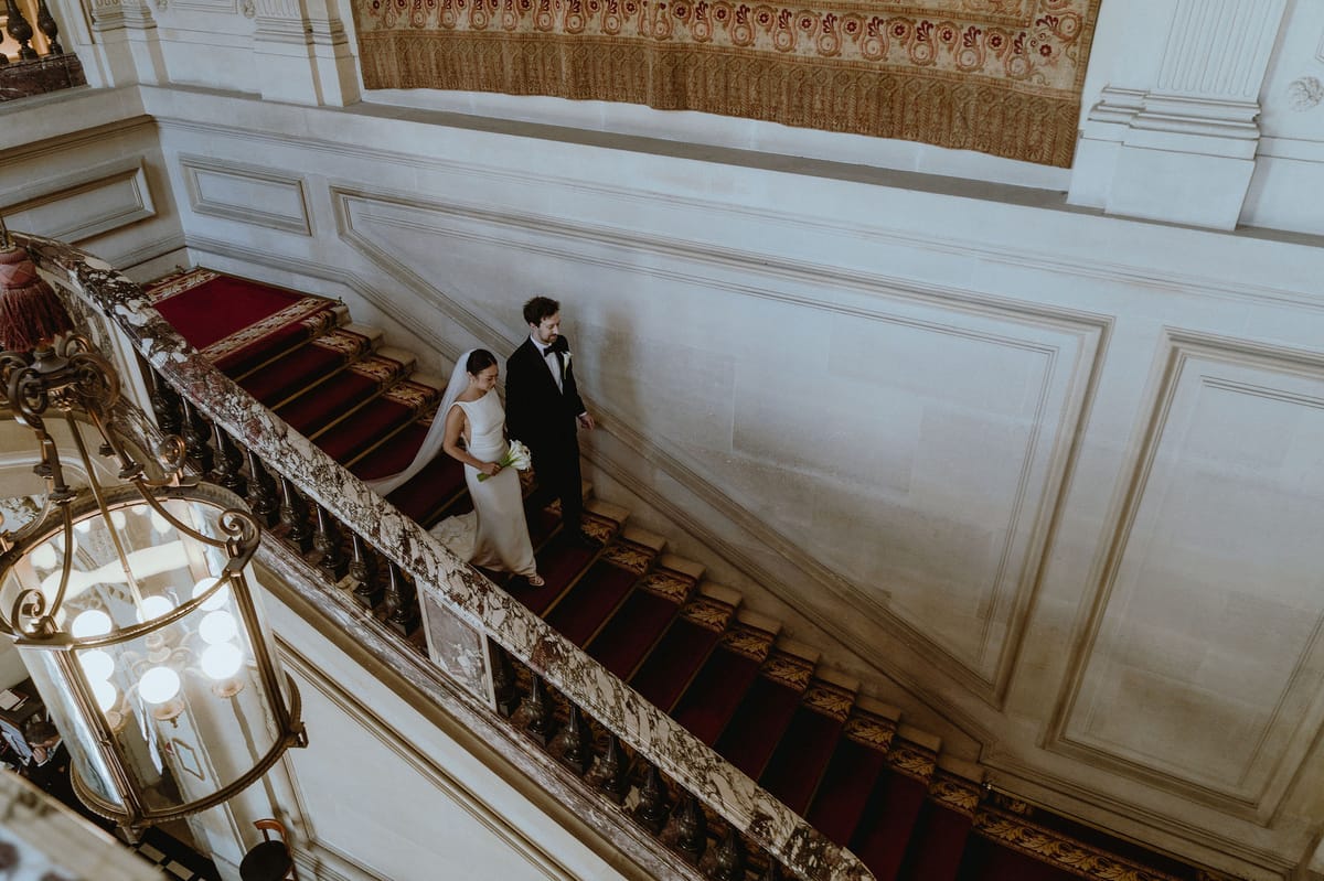 The couple walk down a wide red-carpeted staircase in a grand hall with white panelled walls and a brass lantern.