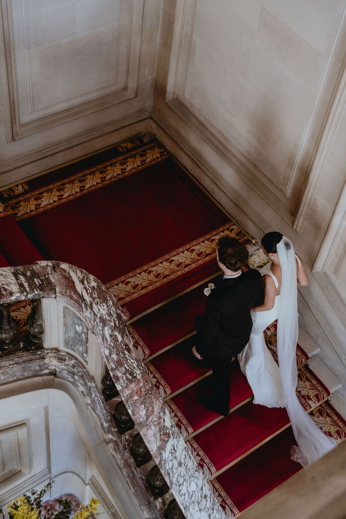 From above, the groom and the bride in a long veil climb a red-carpeted staircase with marble balustrade.