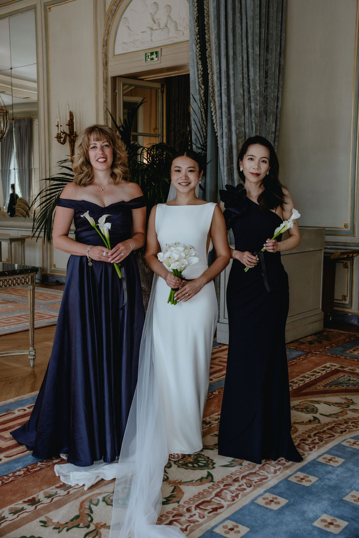 The bride in white stands between two bridesmaids in navy gowns, each holding white flowers, in an ornate room with patterned carpet.