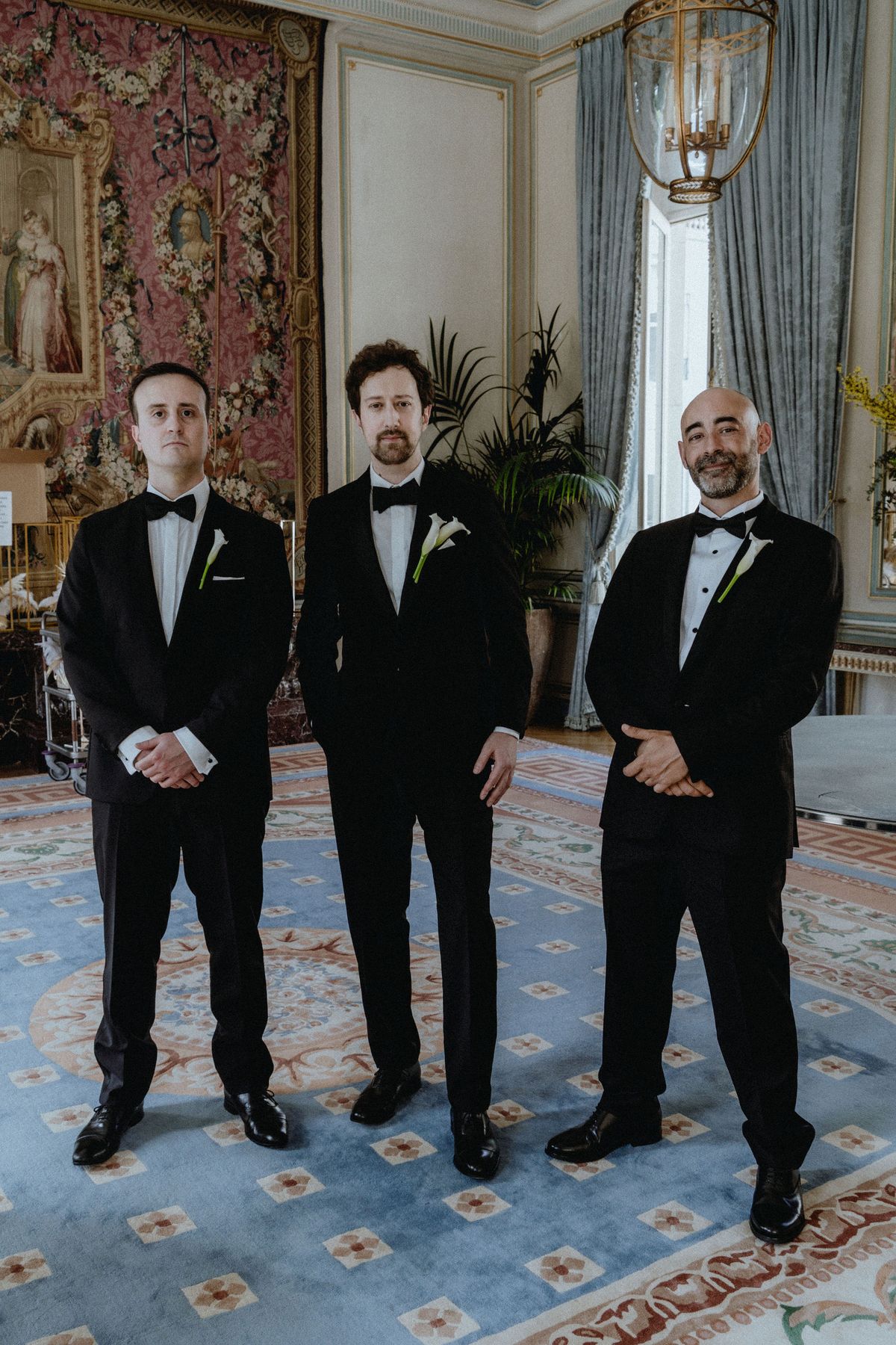 Three men in black tuxedos and bow ties stand in a row on a blue patterned carpet in an ornate room with tapestries.