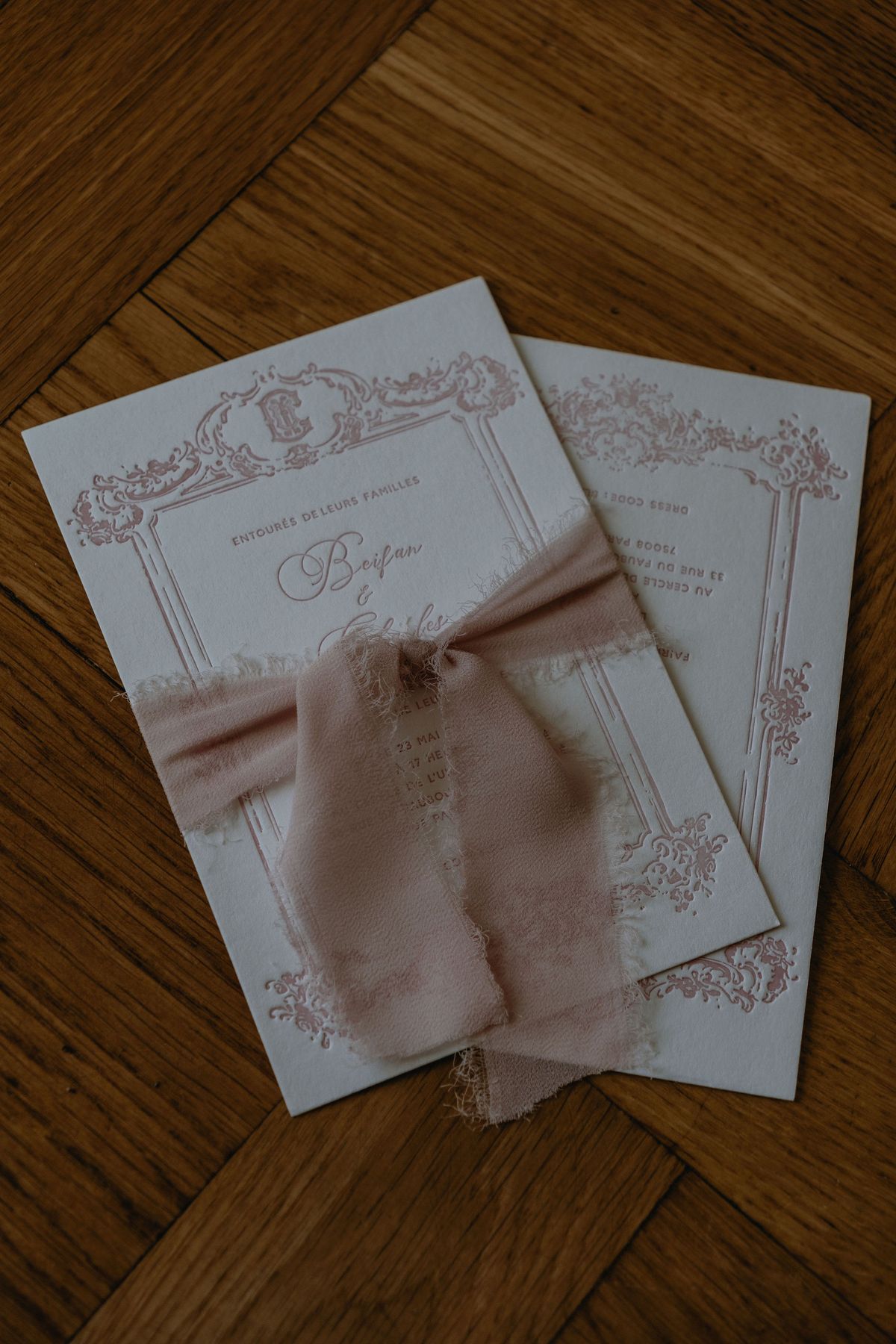 Two printed programmes with delicate pink borders, tied with a blush silk ribbon, lie on a herringbone wooden floor.