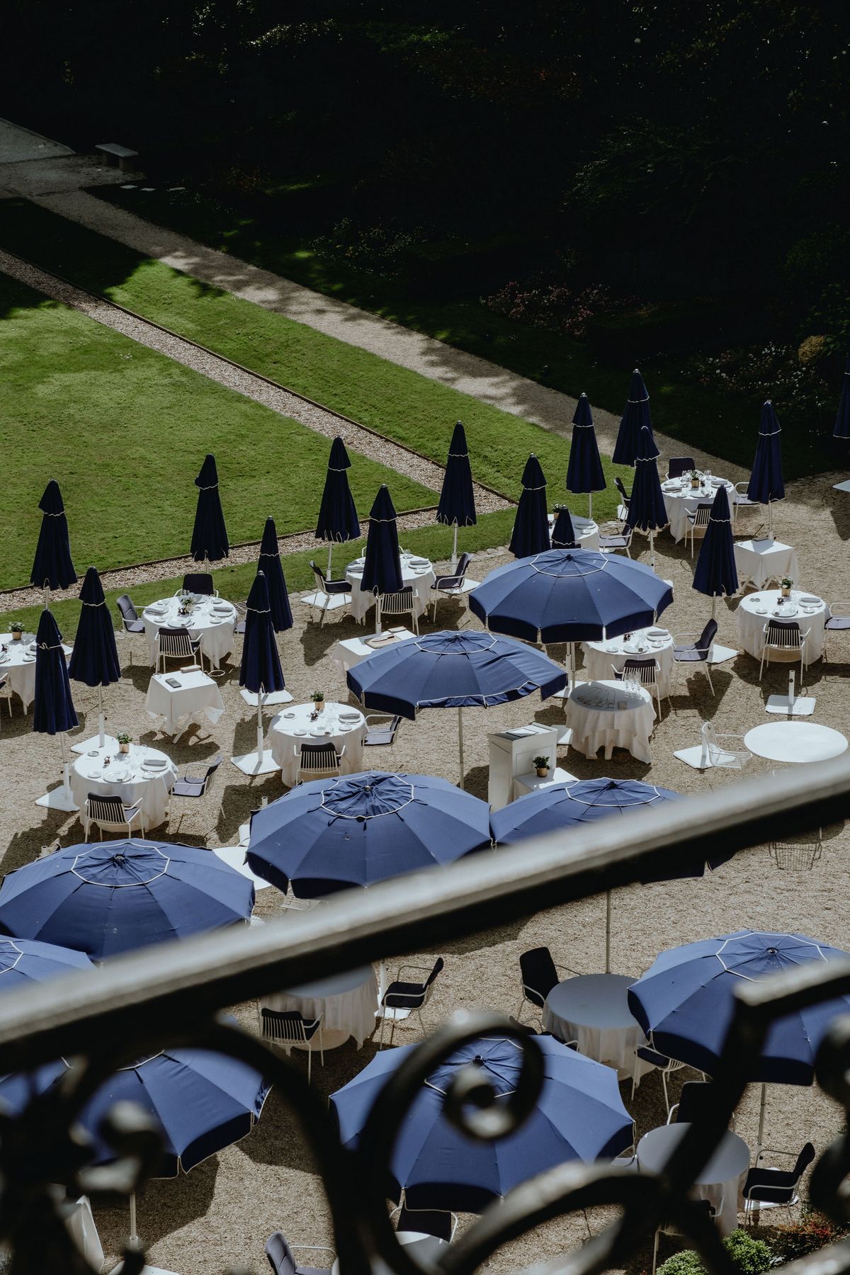 From a balcony, a terrace with tables under navy blue parasols overlooks a green lawn and trimmed hedges.