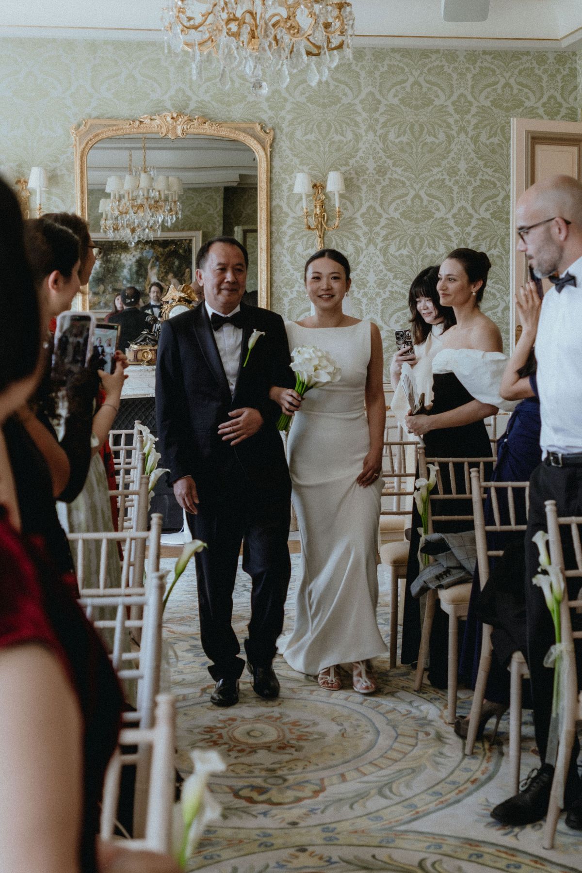 The bride in a simple white gown walks down an aisle between chairs beside a man in a dark suit, while guests look on.
