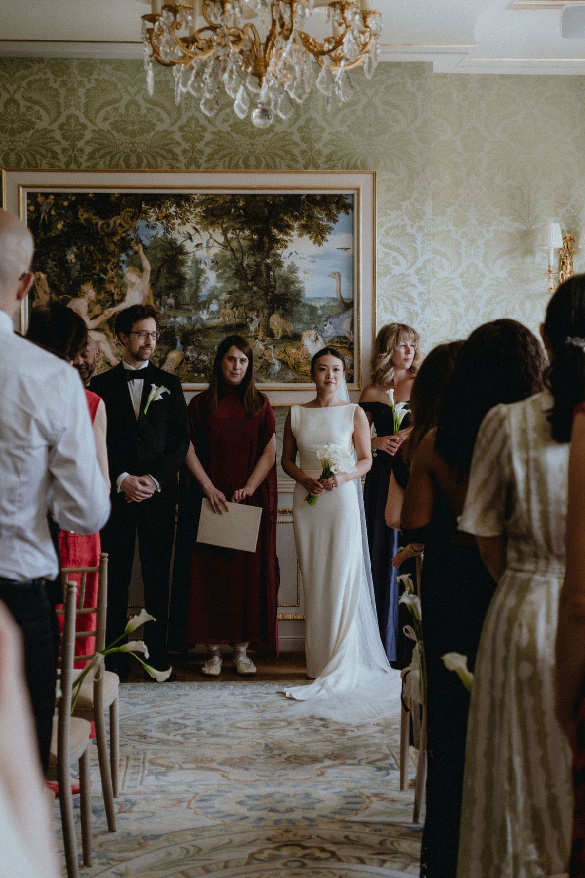 The couple stand at the end of the aisle in front of a painting, surrounded by guests, during the ceremony.