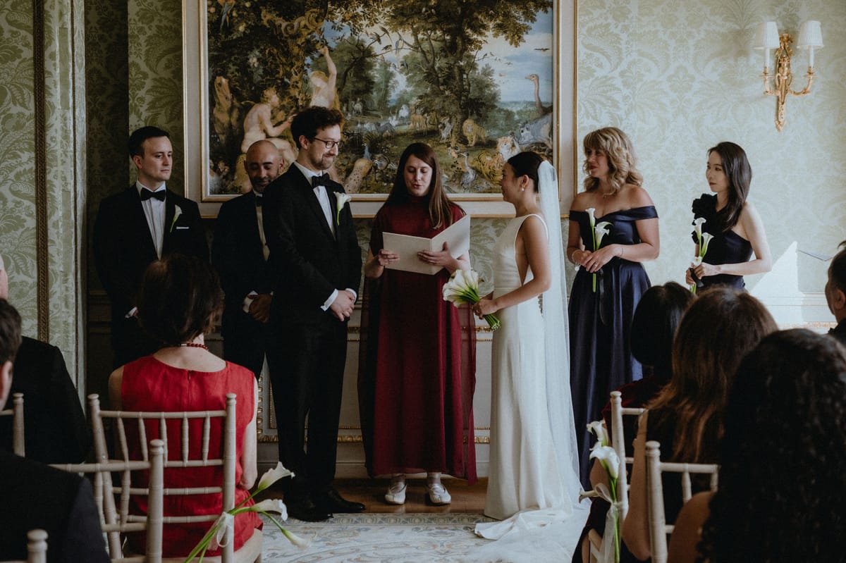 The bride and groom stand facing each other beside an officiant in a red dress, with attendants and seated guests in an ornate room.