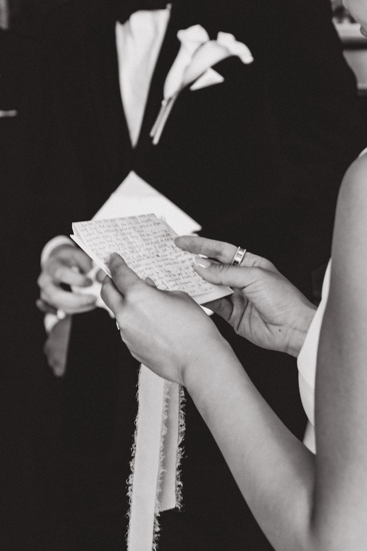 In black and white, hands hold a folded handwritten note tied with ribbon beside a suit lapel with a calla lily.