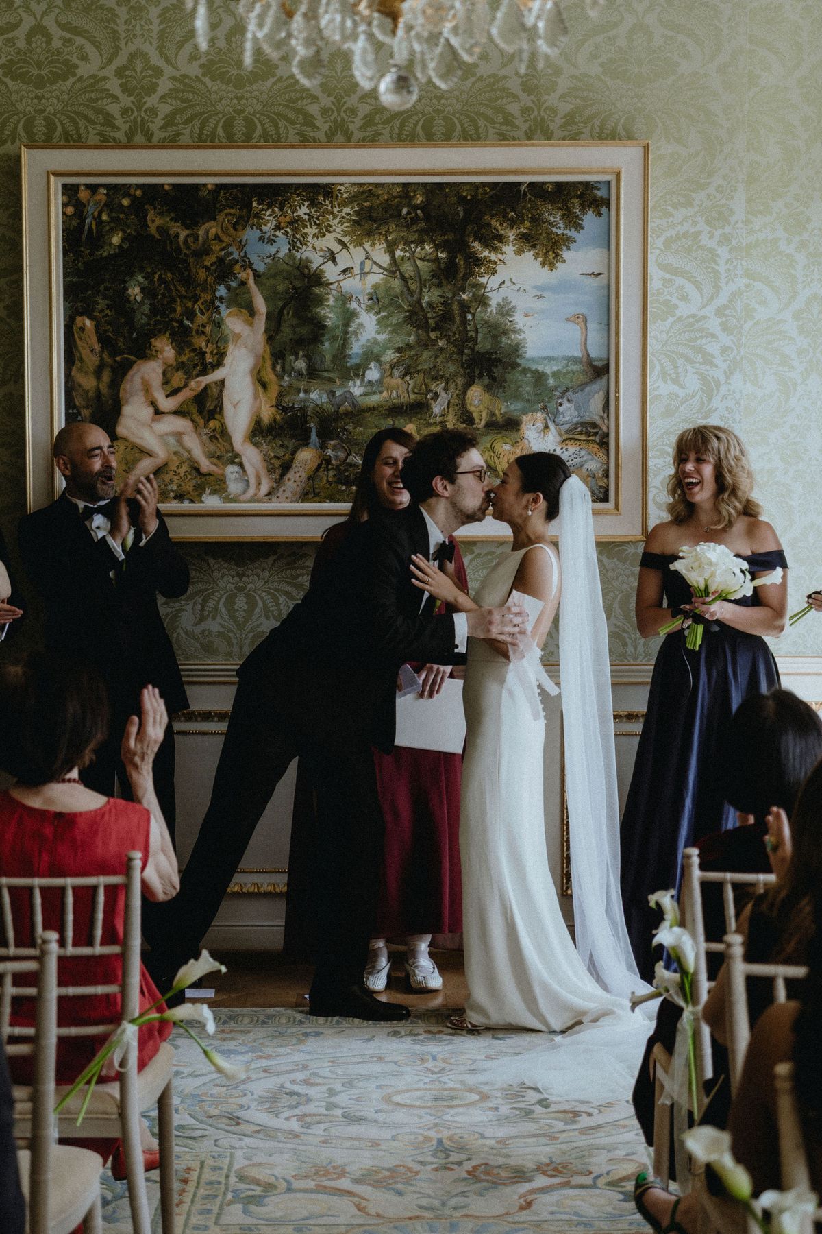 A man in a dark suit kisses the bride on the cheek in front of a large painting, as guests look on.
