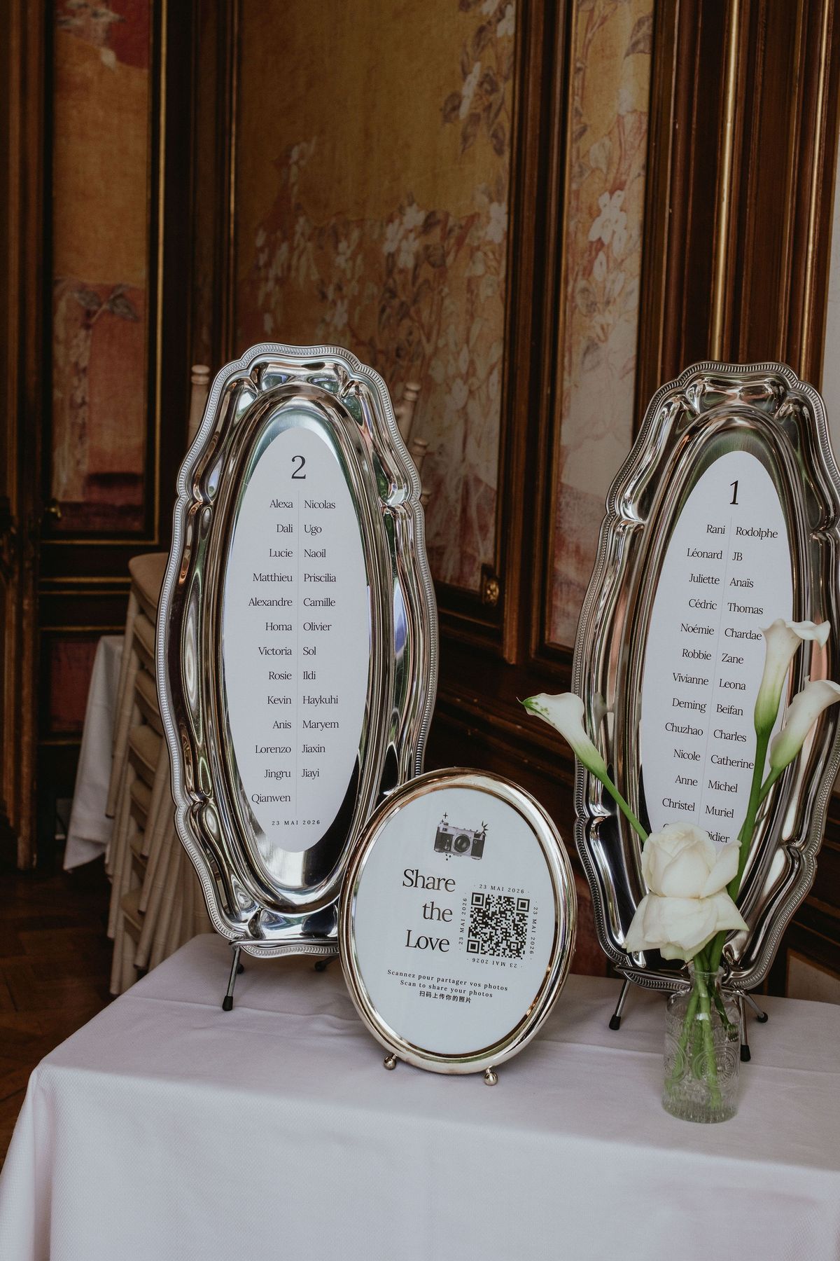 Two oval silver frames hold table lists beside a small framed sign with a QR code and a vase of white flowers.