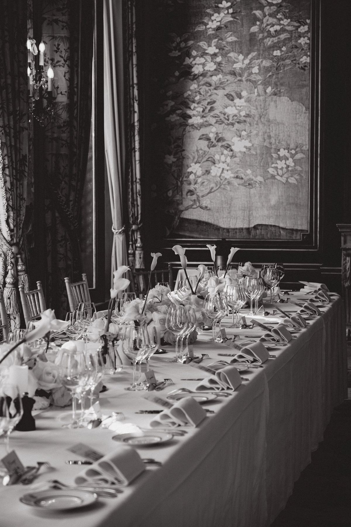 A long table dressed with a cloth, glasses, plates and small flower arrangements stands in a dark panelled room with curtains, in black and white.