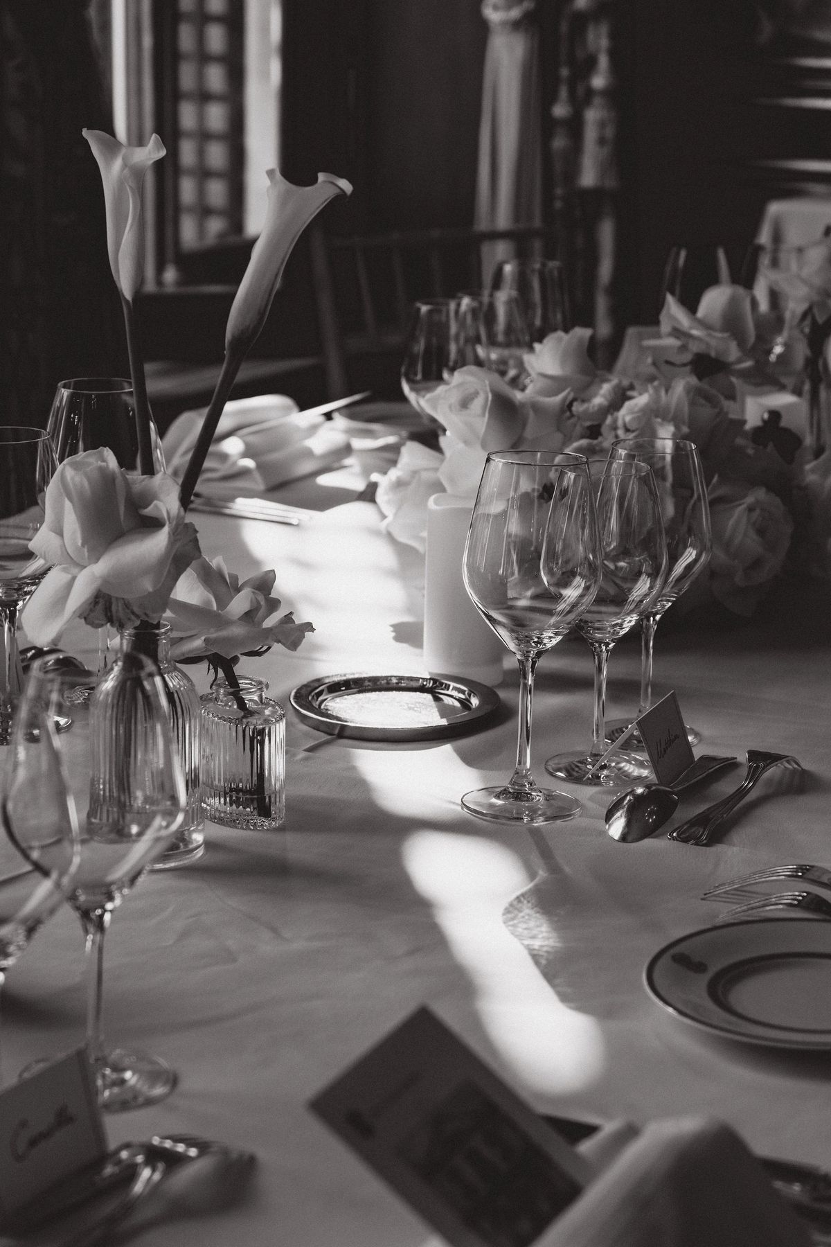 In black and white, sunlight falls across a table set with wine glasses, plates, place cards and tall calla lilies.