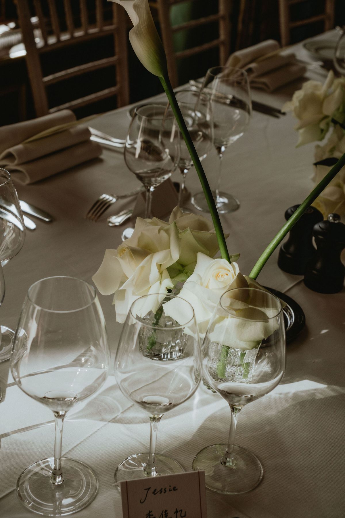 White roses and calla lilies in a small vase sit among wine glasses and a name card on a white tablecloth.