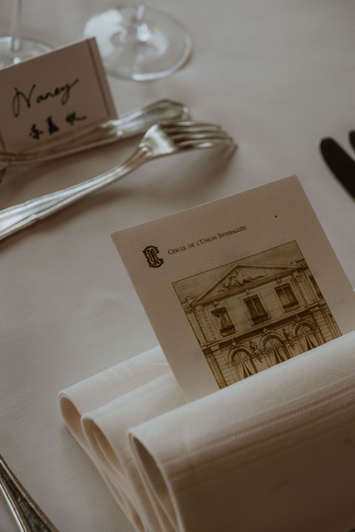 A close view of a table setting with cutlery, a folded napkin, a handwritten name card and a menu card showing a building.