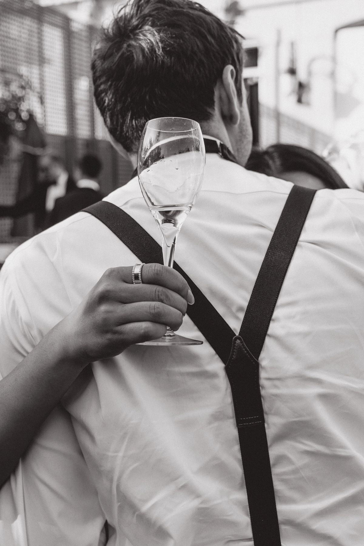 In black and white, a man in a shirt and braces holds a champagne flute while embracing another person.