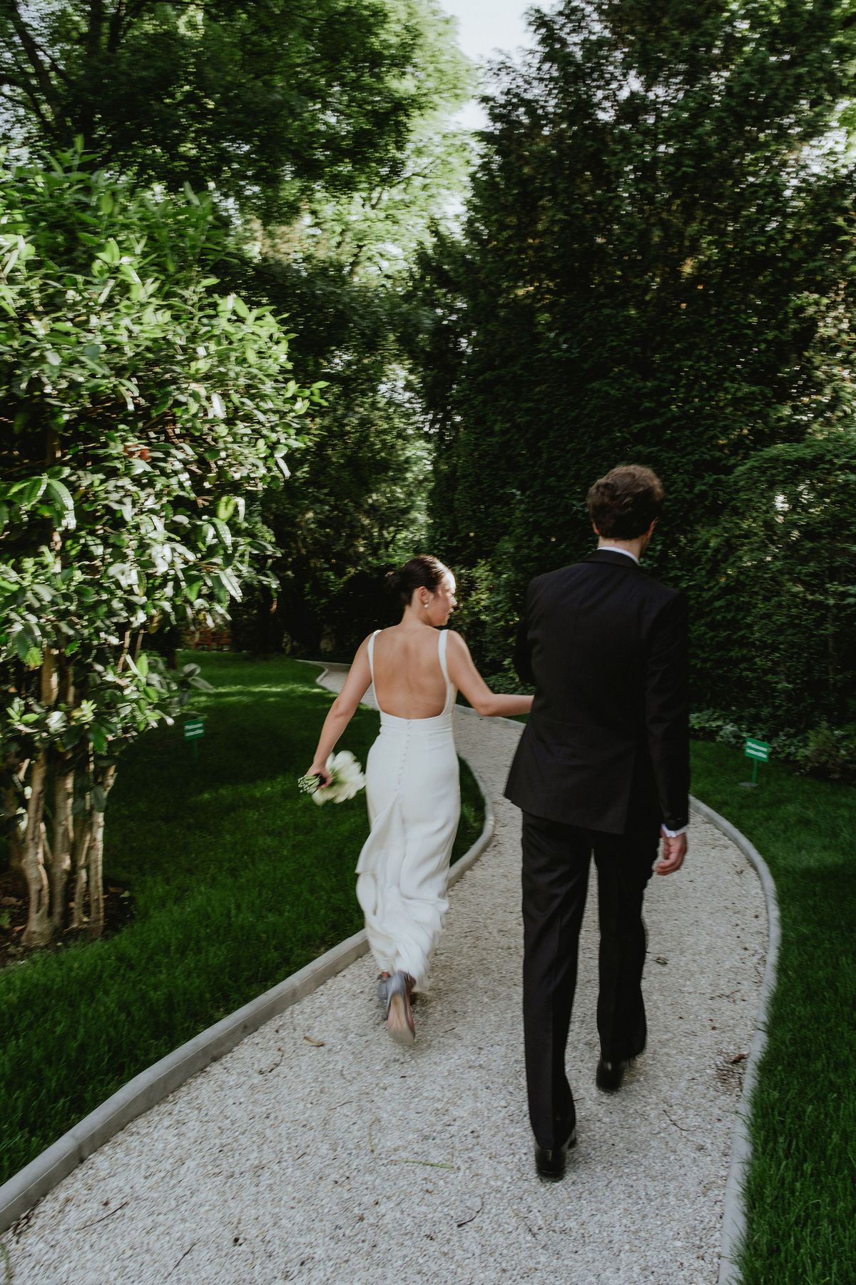 Seen from behind, the bride in a white dress and the groom in a dark suit walk along a curving gravel path between greenery.