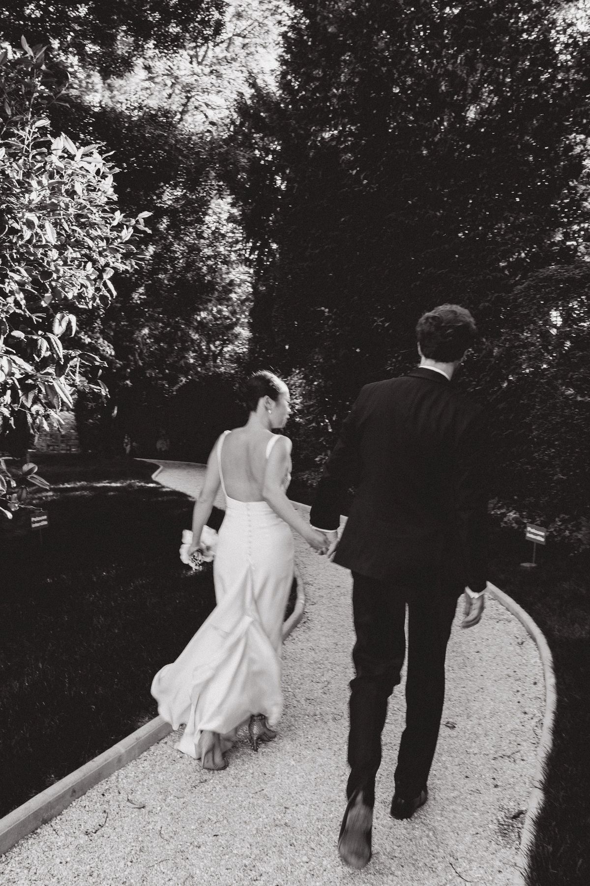 The bride in a long white dress and the groom in a dark suit walk away along a gravel path between trees, in black and white.