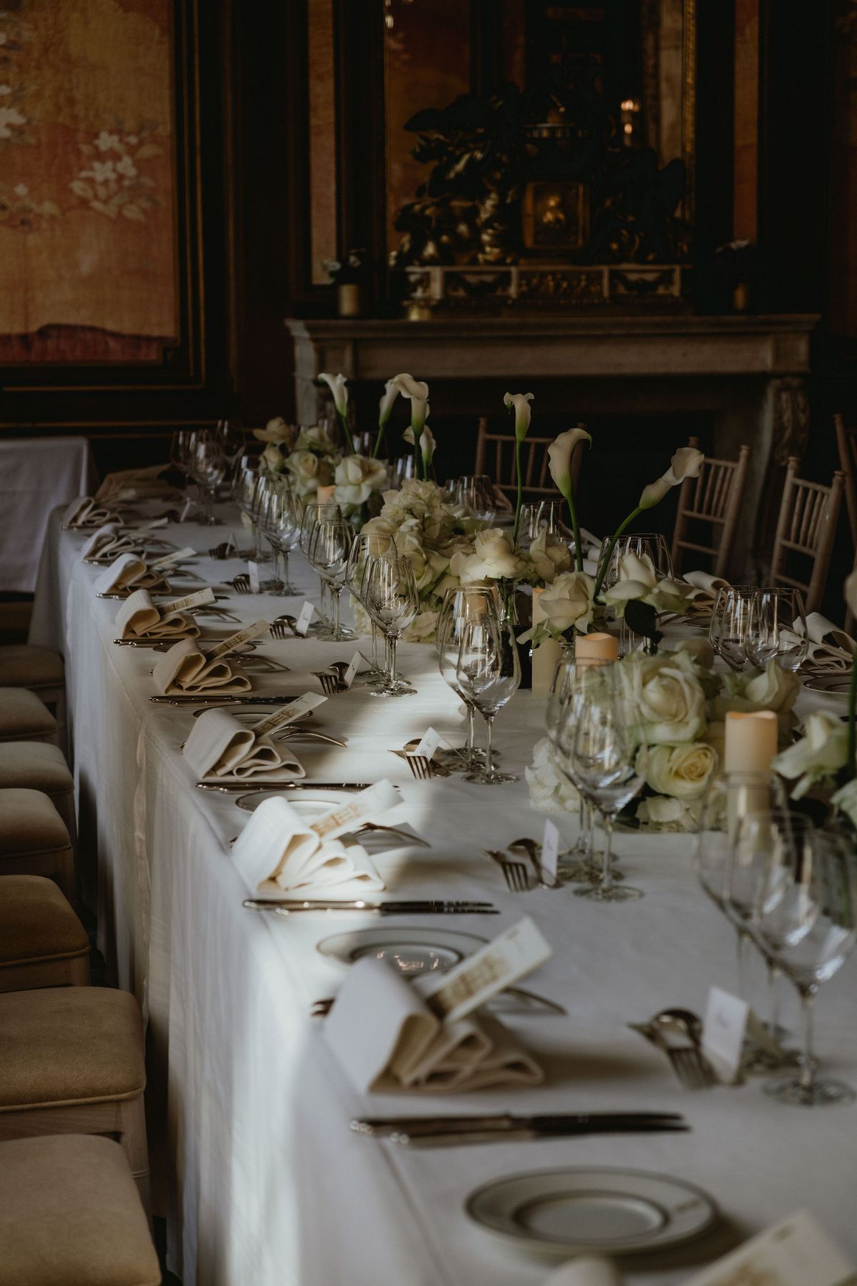 A long table dressed with white flowers, glasses and folded napkins stands in a dark panelled room with a fireplace.