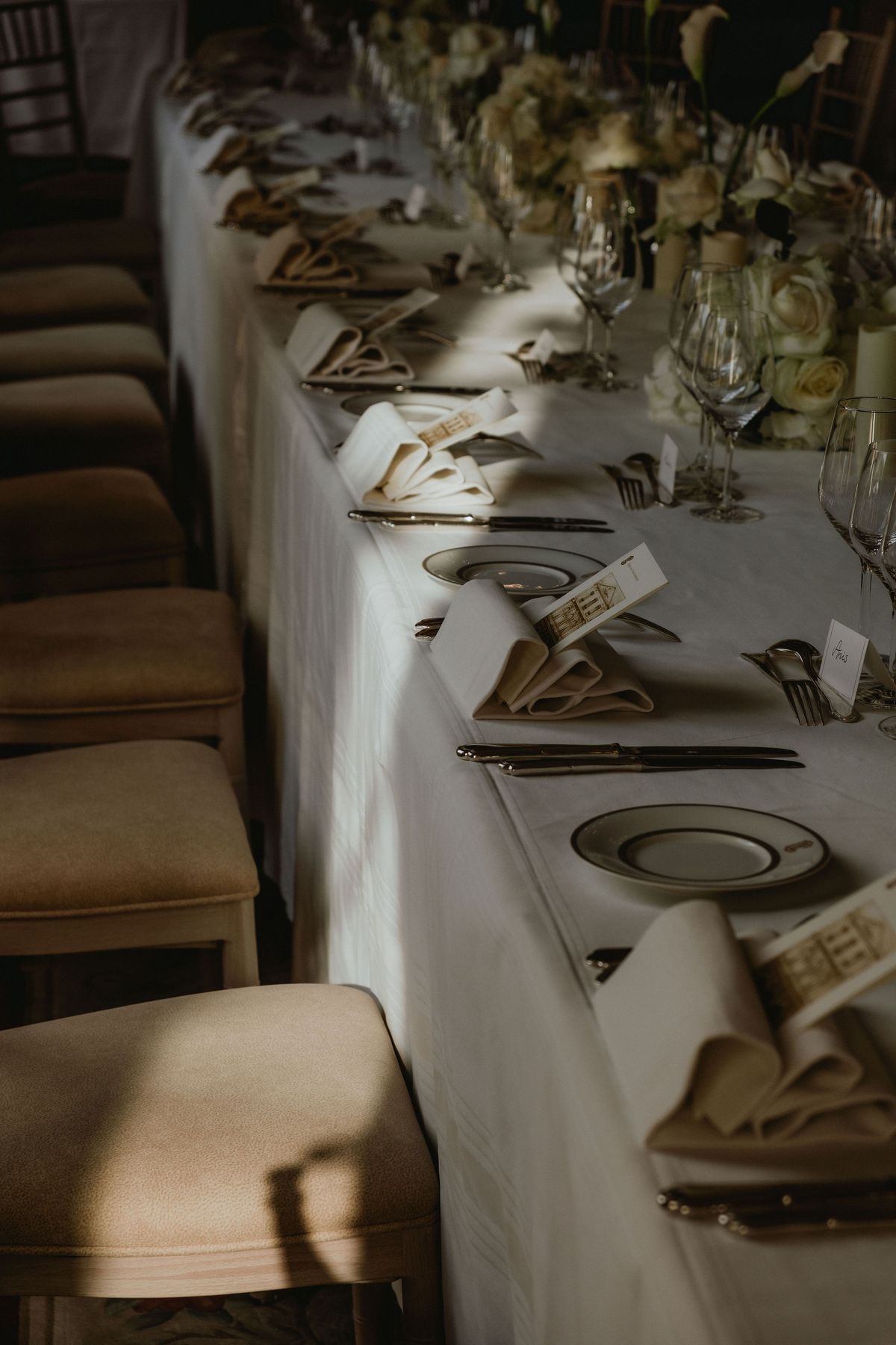 A long table with white linen is laid with plates, glasses, folded napkins and name cards, flanked by upholstered chairs.