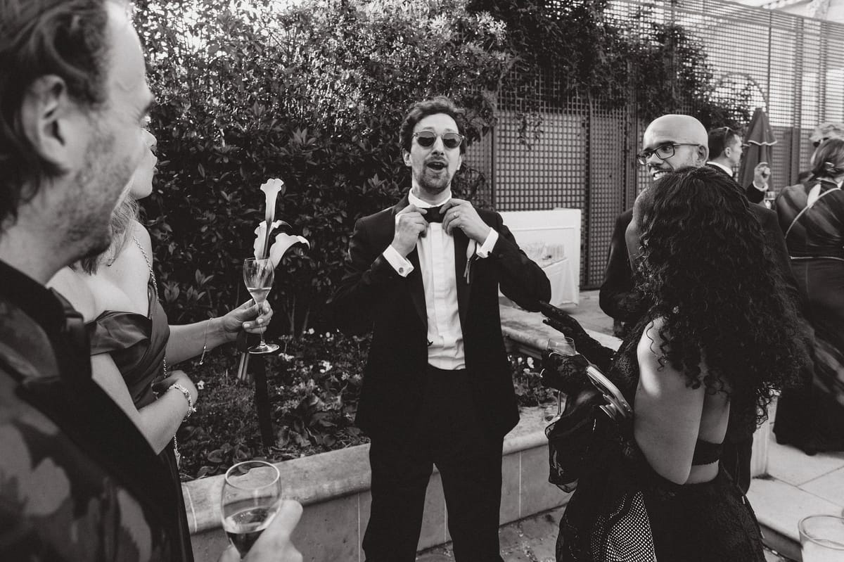 In black and white, the groom in sunglasses adjusts his bow tie while guests holding drinks and a calla lily stand around him.