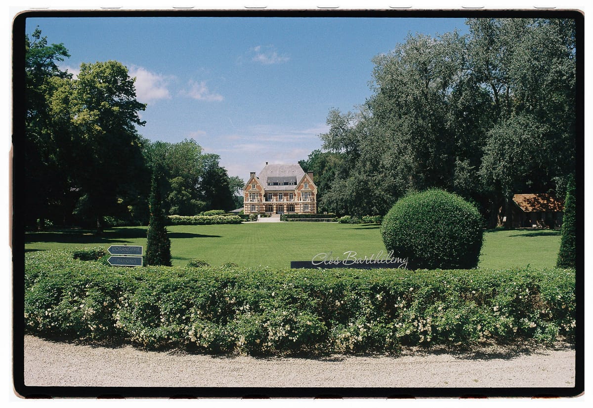 A distant chateau stands beyond a green lawn, a rounded topiary and a low hedge, under a blue sky with tall trees.