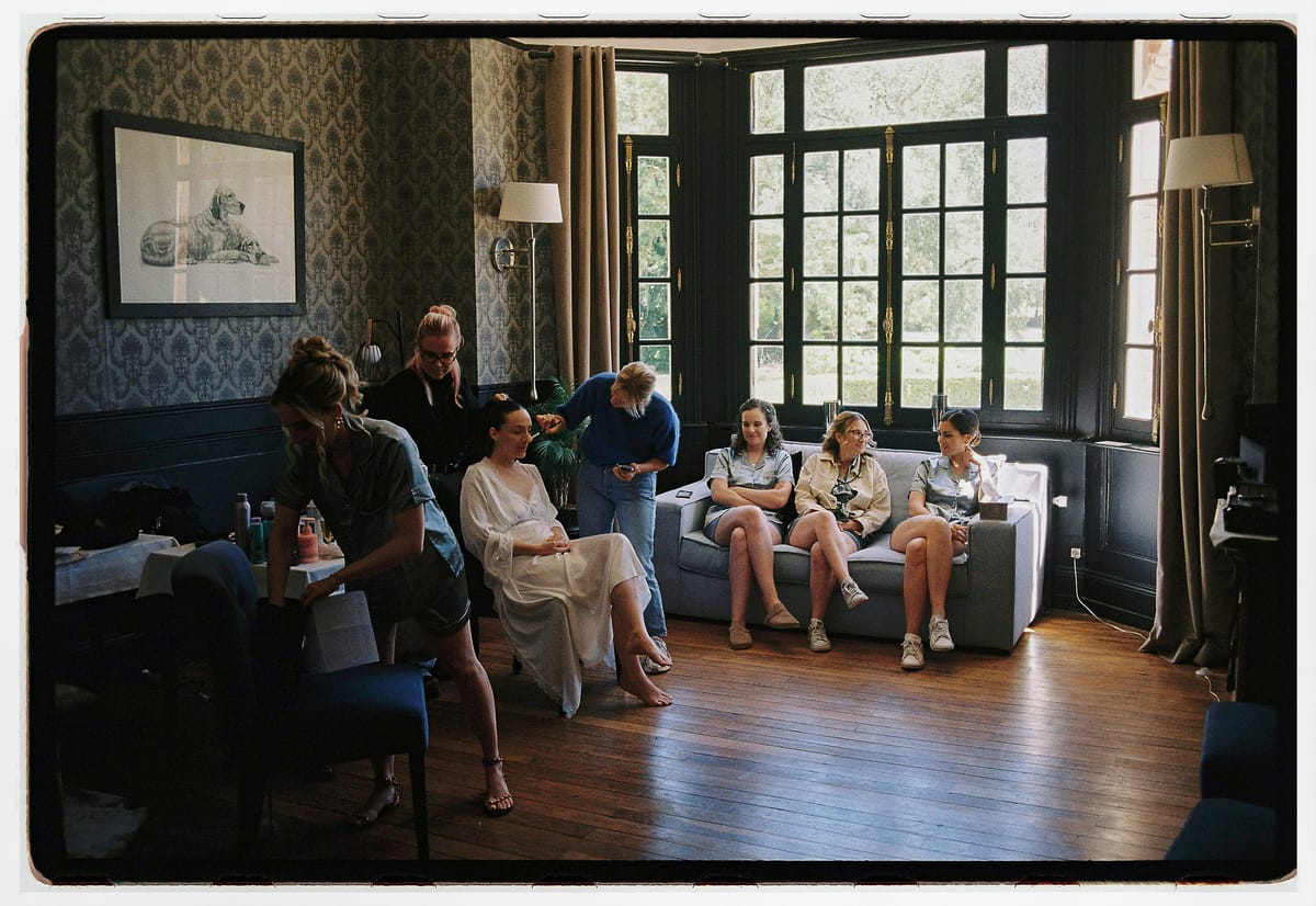 Several people sit and stand in a room with patterned blue walls, tall windows and a wooden floor, some on a sofa.