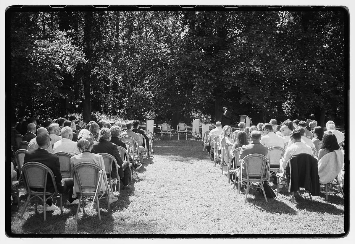 In black and white, guests sit on chairs on both sides of a gravel aisle in a clearing among tall trees.