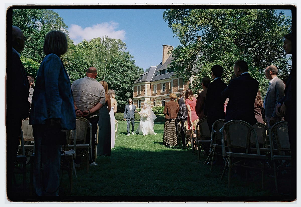 Guests in silhouette watch the couple standing at the end of the aisle during an outdoor ceremony, with a chateau behind the trees.