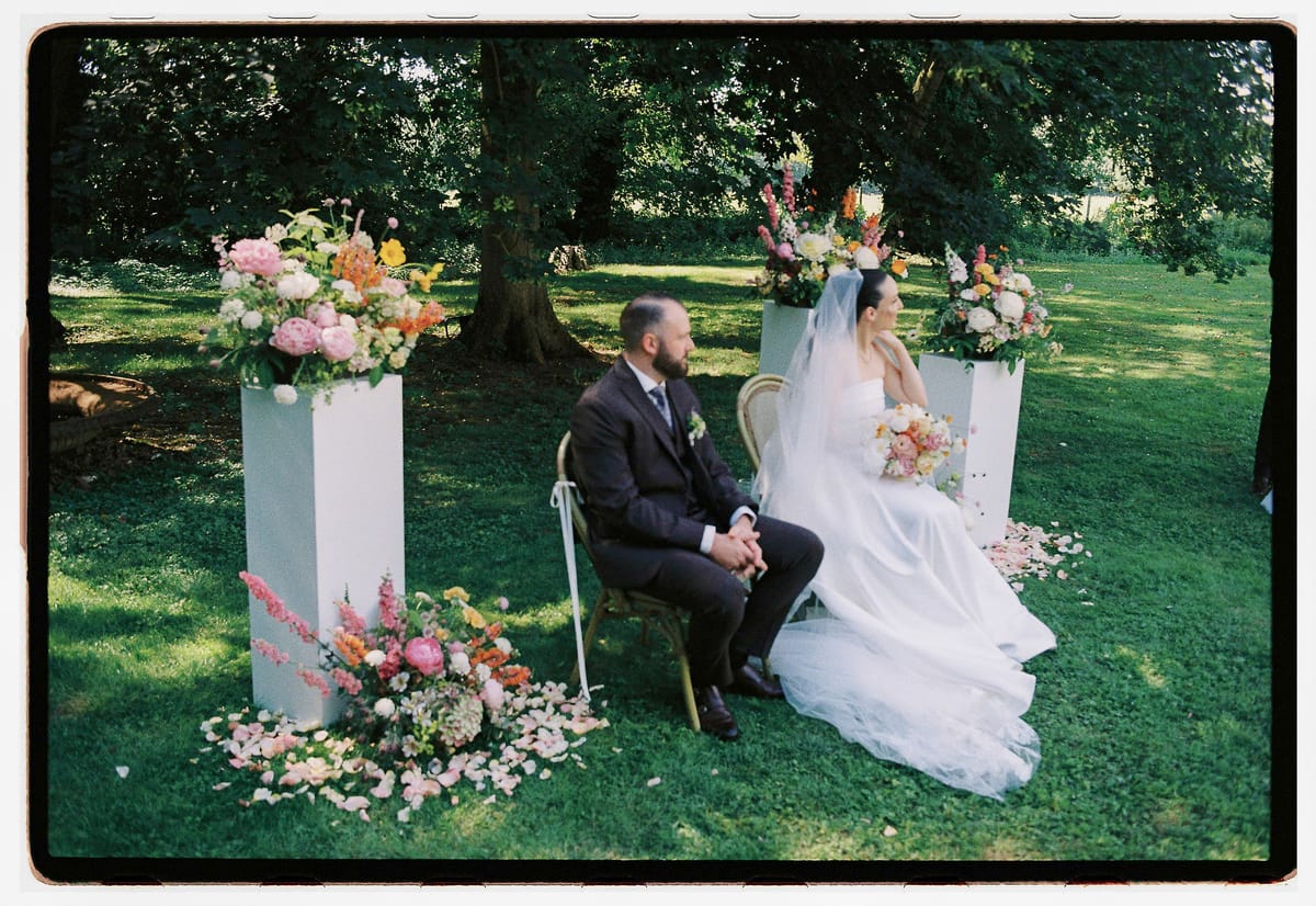 The groom in a dark suit and the bride in a layered white gown sit on chairs between white pillars topped with flowers.