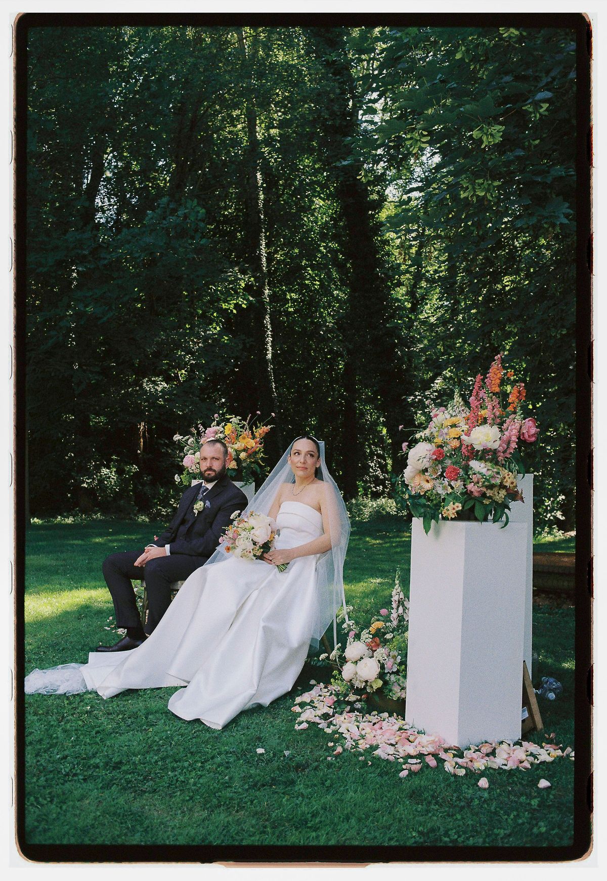 The groom and the bride in a full white gown sit on a lawn beside a white pillar topped with colorful flowers.