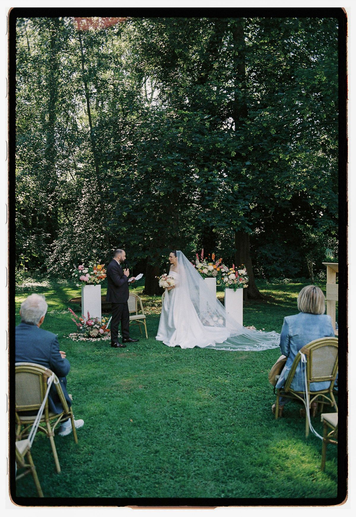 The bride stands facing an officiant between white flower pillars on a lawn, with guests seated on chairs at either side.