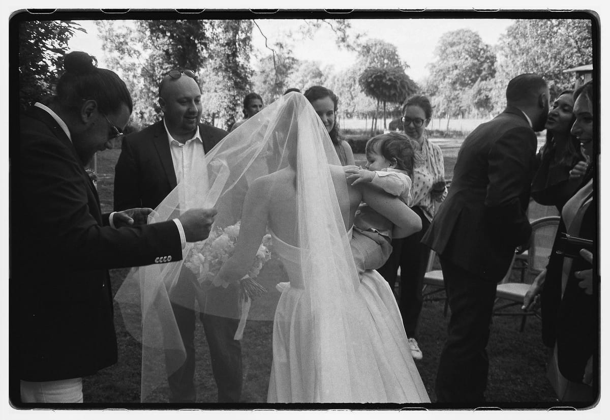 Black and white framed image of guests gathered around the bride in her veil, with several people leaning in and one holding a child.