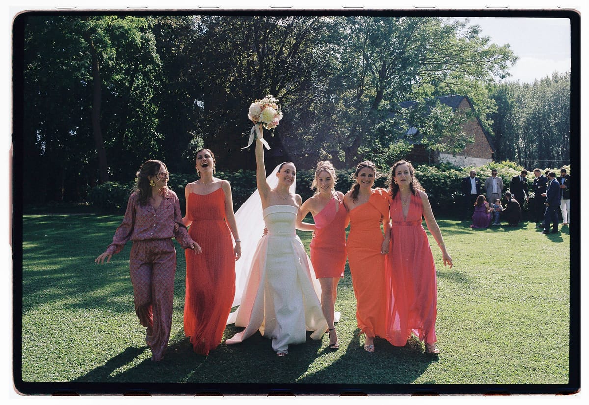 The bride raises her bouquet while walking across a lawn with five bridesmaids in orange and pink outfits, guests behind them.