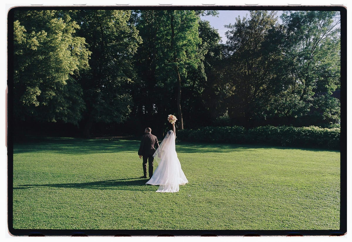 The couple stands small on a large green lawn bordered by tall trees, the bride in a long white gown.