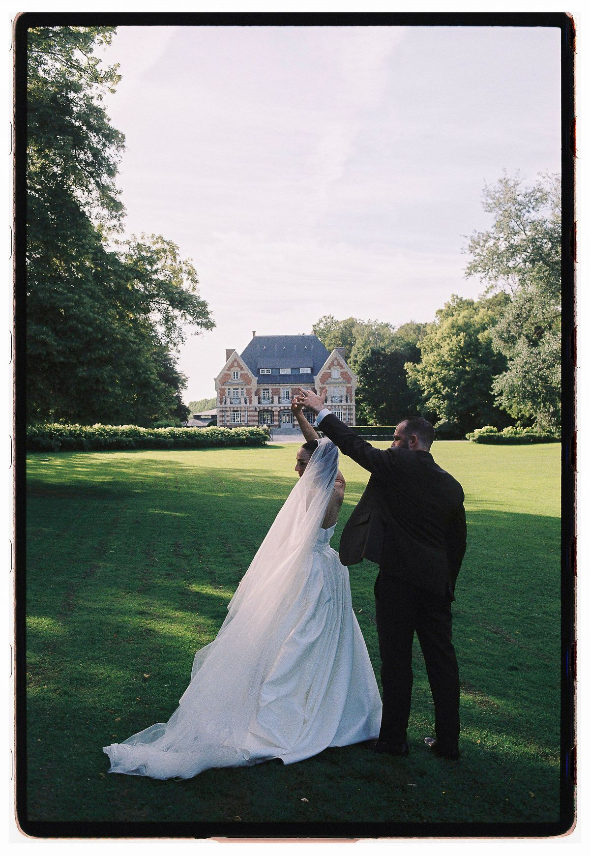 A bride in a long white gown and a groom in a dark suit stand on a lawn, facing a distant chateau.