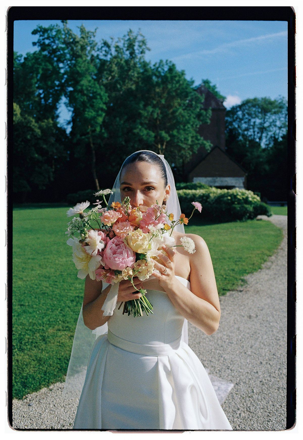 The bride in a white dress and veil holds a large bouquet in front of her face on a lawn beside a gravel path.
