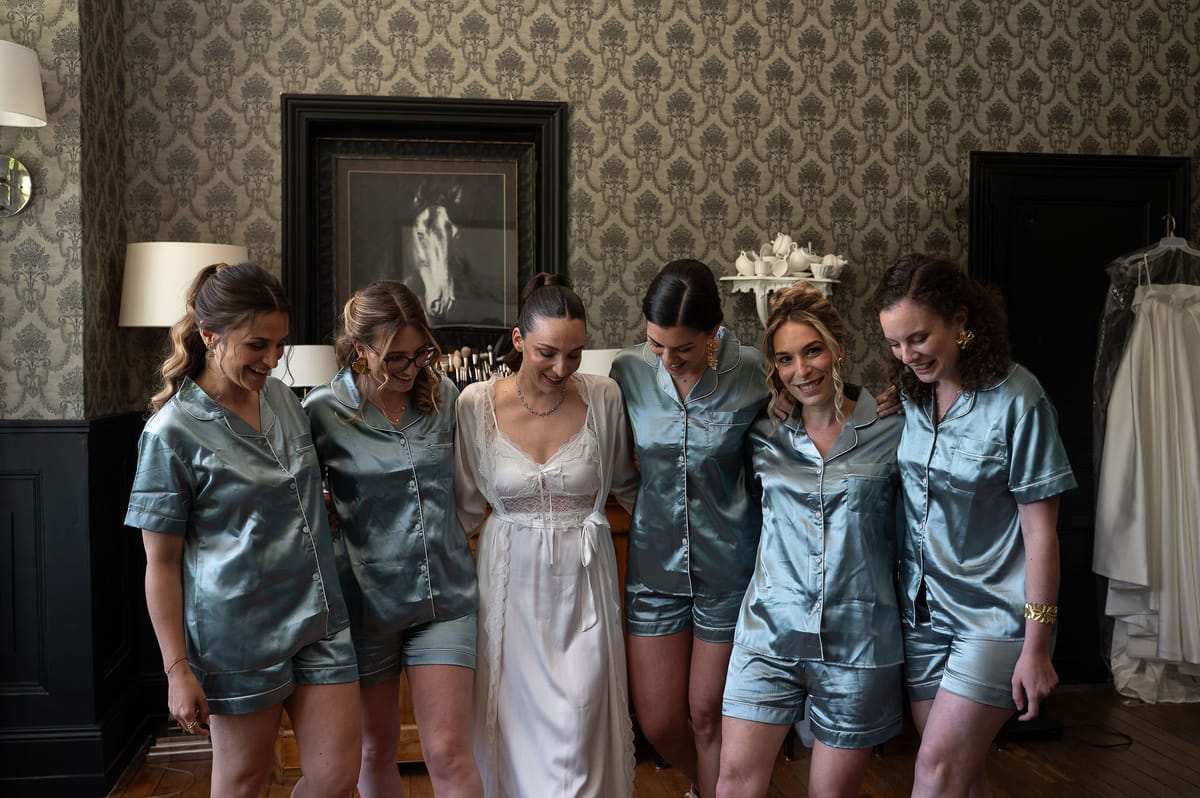Six women stand close together in a patterned-wallpaper room, five in blue satin pajamas and one in a white robe, smiling and looking down.